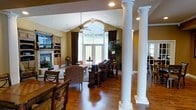 This single-story space showcases a thoughtfully designed open-concept layout that seamlessly connects the dining and living areas. Stately white fluted columns with classical detailing serve as elegant architectural dividers, defining each zone while maintaining a sense of openness throughout. The vaulted ceiling adds vertical drama and an airy quality to the living area, which is anchored by a ceiling fan and recessed lighting.

A prominent arched window flanked by full-length glass French doors floods the space with natural light, framing views of the outdoors and creating an inviting focal point. Rich, dark hardwood flooring runs continuously through the space, complementing the warm golden-beige wall tones and the dark-stained wood furnishings. A built-in entertainment center and fireplace surround occupy one wall of the living area, adding both function and character.

The dining area, positioned near the columned entry, features traditional wood furniture that echoes the warm, classic aesthetic carried throughout the space. Overall, the interior exudes a traditional yet welcoming ambiance with refined architectural details.