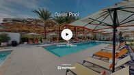 Located at 14147 Darwin Drive in Moreno Valley, California, this single-level outdoor amenity space offers a resort-inspired pool experience set against a dramatic natural landscape. The centerpiece is a generously sized, crystal-clear swimming pool with a dedicated spa, both finished with vibrant blue water that contrasts beautifully with the sun-drenched concrete surround. An expansive pool deck provides ample space for relaxation, furnished with rows of orange and gray lounge chairs and a variety of market and cantilever umbrellas in coordinating tones. Towering palm trees are planted throughout the deck, lending a tropical ambiance and natural shade to the space. The perimeter of the area is thoughtfully landscaped with shrubs, ornamental grasses, and young trees, creating a lush border between the deck and the surrounding community. The backdrop is defined by a striking hillside of rugged boulders and golden chaparral, offering a quintessential Southern California desert-meets-oasis aesthetic. Glimpses of the adjacent Mediterranean-style residential buildings — characterized by white stucco exteriors and clay tile rooflines — further frame the setting. The overall atmosphere is bright, open, and inviting, designed to encourage outdoor recreation and leisurely enjoyment under the wide Inland Empire sky.