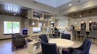 This single-floor space showcases a thoughtfully designed open-concept layout that seamlessly integrates multiple functional areas within one cohesive environment. A striking freestanding geometric bookshelf unit serves as the central architectural element, acting as a stylish room divider that defines separate zones without fully enclosing them, maintaining an airy, connected feel throughout.

The lounge area features a sleek linear fireplace set against a decorative wall, complemented by contemporary seating arranged over a richly patterned area rug. Adjacent to this is a dedicated workspace equipped with ergonomic office chairs and workstations, offering a professional yet comfortable setting. A round white dining or meeting table surrounded by navy upholstered chairs anchors another functional zone, ideal for collaborative gatherings.

Toward the rear, a well-appointed kitchenette or refreshment station is visible, featuring white cabinetry, a decorative tile backsplash, and beverage equipment. The ceiling design incorporates varied levels and finishes, including recessed lighting and wood-paneled sections, adding depth and visual interest. The overall ambiance is modern and polished, balancing functionality with refined aesthetic detail.