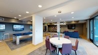 This single-floor space is designed as a multifunctional communal area that seamlessly integrates several distinct zones within an open-plan layout. The interior features light hardwood flooring and smooth white ceilings equipped with recessed lighting throughout, creating a bright and welcoming atmosphere. A circular pendant light fixture serves as a focal point above a round white dining table surrounded by plum-toned upholstered chairs, anchored by a textured area rug. Adjacent to the dining area, a lounge zone is furnished with a deep blue sectional sofa accented with red and blue throw pillows, positioned near a wall-mounted flat-screen television. A billiards table occupies a dedicated entertainment section toward the rear of the space, accompanied by additional seating and another mounted display screen. Structural columns are incorporated into the design as clean architectural elements that help define the different zones without fully enclosing them. The overall ambiance is contemporary and polished, with a cohesive color palette of blues, grays, and warm wood tones that lends the space a sophisticated yet comfortable character suited for social gatherings and recreation.
