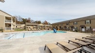 Located at 1864 Brevard Rd in Arden, North Carolina, this outdoor amenity area offers a well-appointed community pool and lounge space designed for relaxation and social gathering. The centerpiece is a generously sized freeform swimming pool surrounded by a broad concrete deck furnished with cushioned lounge chairs, ideal for sunbathing and leisure. The pool area is enclosed by a wrought iron fence, providing both security and an open, airy feel with views of the surrounding autumn-tinged tree line and multi-story residential buildings finished in warm tan siding with dark shutters.

Adjacent to the pool is a covered outdoor patio pavilion that serves as a true gathering space. This area features a wood-paneled vaulted ceiling with ceiling fans, a mounted flat-screen television, a brick wood-burning fireplace, and comfortable seating arrangements including sofas and dining tables with chairs — perfect for entertaining in any weather. Outdoor lighting and a pergola accent add architectural character to the space.

The complex also includes an outdoor grill station, reinforcing the area's focus on community living. The building exteriors reflect a traditional design with brick accents, complementing the natural landscape of the western North Carolina setting. Safety features such as an emergency phone are prominently placed throughout the grounds.