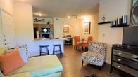 This single-story space showcases an open-concept floor plan that integrates the kitchen, dining, and living areas into one cohesive environment. The kitchen features dark wood cabinetry, black appliances including a refrigerator and range, and a breakfast bar with granite-style countertops, accommodated by two bar stools for casual seating. Track lighting illuminates the kitchen area, complementing the flush-mount ceiling fixtures throughout. An arched passageway adds an architectural detail that subtly defines the transition between spaces. The dining area accommodates a round glass-top table surrounded by vibrant orange upholstered chairs, creating a bold focal point. A built-in desk nook is tucked nearby, offering a compact workspace. The living area features rich, dark hardwood-style flooring that runs throughout, anchoring a zebra-print accent chair and an ottoman. Warm, neutral wall tones create an inviting ambiance, while framed artwork adds decorative character. The open layout promotes a sense of spaciousness and easy flow between functional zones.