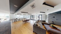 This thoughtfully designed single-floor amenity space offers a seamless blend of comfort and functionality across several well-defined zones. The expansive open-plan layout is anchored by warm, light-toned hardwood flooring that flows throughout, creating a cohesive and inviting atmosphere.

The main gathering area features a generous sectional sofa and a large dining table with modern chairs, perfect for group socializing or casual meetings. Floor-to-ceiling glass sliding doors and full-height windows flood the interior with abundant natural light while providing direct access to an outdoor terrace, where additional seating is visible beyond.

A dedicated media lounge area is highlighted by a dramatic deep-teal geometric accent wall with a large wall-mounted flat-screen television, complemented by plush seating and a low-profile ottoman. A quiet nook with wraparound windows offers elevated views of the surrounding neighborhood and waterway, furnished with comfortable lounge chairs ideal for relaxed reading or conversation.

Along one wall, a sleek bar counter with dark countertops and wood barstools serves as a social hub, backed by a gallery of bold abstract artwork on a deep navy feature wall. An additional bar-height counter with integrated cabinetry provides practical workspace. The space is finished with modern track and recessed lighting, a two-tone ceiling design, and refined architectural details throughout.