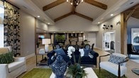 Located at 3701 Governors Club Blvd in Columbus, Ohio, this sophisticated single-story clubhouse is designed to impress with its refined architectural details and upscale amenities. The centerpiece of the space is a soaring vaulted ceiling accented by rich exposed wooden beams and white shiplap paneling, creating a warm yet grand atmosphere. A statement wrought-iron orb chandelier anchors the ceiling, adding an industrial-chic touch to the otherwise classic design. The main lounge area is furnished with plush navy blue velvet sofas and complementary accent chairs arranged around a sleek coffee table, offering a comfortable and stylish gathering space. Lush greenery, fresh floral arrangements, and decorative ceramic accents add life and texture throughout. Moving deeper into the space, a dedicated dining area features elegant tufted seating around dark-finished tables, ideal for social gatherings. A full bar and kitchen area is visible in the background, complete with pendant lighting, dark cabinetry, and professional-grade appliances, making it well-suited for entertaining. Light wood-toned flooring flows seamlessly throughout, tying all areas together. Natural light filters in through large windows and glass doors, enhancing the bright and welcoming ambiance of this well-appointed community clubhouse.