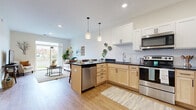 This single-floor residence is designed with a clean, contemporary aesthetic that emphasizes an open and functional layout. The kitchen serves as a central hub, featuring a two-tone cabinet design that pairs natural wood lower cabinets with crisp white upper cabinetry, all complemented by dark countertops and a classic subway tile backsplash. A full suite of stainless steel appliances—including a refrigerator, range, microwave, and dishwasher—adds a polished, modern finish. Pendant lighting above the peninsula and recessed ceiling lights enhance the kitchen's bright, welcoming atmosphere. The kitchen flows seamlessly into the living area, where light wood-style flooring continues throughout. The living room benefits from a large sliding glass door that fills the space with natural light and provides direct access to a private patio overlooking a landscaped outdoor setting. The two bedrooms are carpeted for comfort, each featuring windows that allow ample natural light and views of the surrounding greenery. The bathroom includes a clean vanity with light wood cabinetry, a white countertop, and a shower-tub combination. Throughout the residence, neutral tones and modern finishes create a cohesive, move-in-ready environment.