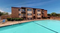 This multi-story building is constructed with classic brick exteriors accented by dark brown trim, lending a sturdy and traditional aesthetic to the structure. The facade is punctuated by multiple large windows and private balconies on the upper floors, offering residents outdoor access and views of the surrounding area. The building spans several floors, providing a substantial and well-proportioned presence.

A standout feature of this property is the expansive outdoor swimming pool situated directly in front of the building. The pool is generously sized, filled with clear turquoise water, and finished with a clean concrete surround that provides ample space for lounging. A wrought iron fence with a secure gate encloses the pool area, clearly delineating it as a dedicated amenity space. Metal pool entry ladders are visible, underscoring the pool's accessibility.

Neatly maintained shrubbery lines the base of the building between the fence and the structure, adding a touch of greenery to the hardscape. The overall setting is bright and open, benefiting from abundant natural sunlight. The combination of the classic brick architecture, private balconies, and resort-style pool creates a functional and visually cohesive outdoor environment.