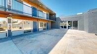 Located at 609 S Thornton Ave in Dalton, Georgia, this modern property presents a thoughtfully designed multi-unit complex centered around a generous open-air courtyard. The building's exterior showcases a striking combination of painted gray brick, warm wood-paneled soffits, and bold blue entry doors, creating a contemporary aesthetic that sets it apart. Ground-floor units feature large glass sliding doors and windows that open directly onto the expansive concrete courtyard, while upper-level units are accessible via an exterior corridor lined with sleek black metal railings, offering an open-air walkway overlooking the common area below. The courtyard serves as the heart of the property, providing ample space for outdoor living and gathering. It is furnished with comfortable seating arrangements, including lounge chairs and accent tables, ideal for relaxing or socializing. A dedicated grilling station is built into the perimeter wall, accompanied by additional seating and a patio table, making it well-suited for outdoor dining. The boundary walls feature decorative cinder block detailing and raised wooden planter beds, adding texture and greenery to the space. Under clear open skies, the courtyard benefits from abundant natural light throughout the day, enhancing the bright and welcoming atmosphere of this well-appointed property.
