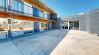 Located at 609 S Thornton Ave in Dalton, Georgia, this modern property presents a thoughtfully designed multi-unit complex centered around a generous open-air courtyard. The building's exterior showcases a striking combination of painted gray brick, warm wood-paneled soffits, and bold blue entry doors, creating a contemporary aesthetic that sets it apart. Ground-floor units feature large glass sliding doors and windows that open directly onto the expansive concrete courtyard, while upper-level units are accessible via an exterior corridor lined with sleek black metal railings, offering an open-air walkway overlooking the common area below. The courtyard serves as the heart of the property, providing ample space for outdoor living and gathering. It is furnished with comfortable seating arrangements, including lounge chairs and accent tables, ideal for relaxing or socializing. A dedicated grilling station is built into the perimeter wall, accompanied by additional seating and a patio table, making it well-suited for outdoor dining. The boundary walls feature decorative cinder block detailing and raised wooden planter beds, adding texture and greenery to the space. Under clear open skies, the courtyard benefits from abundant natural light throughout the day, enhancing the bright and welcoming atmosphere of this well-appointed property.