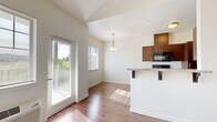 This thoughtfully designed one-bedroom, one-bathroom residence offers a functional and modern single-floor layout. The galley-style kitchen is a standout feature, outfitted with rich dark wood cabinetry, granite countertops, black stainless steel appliances including a range, microwave, refrigerator, and dishwasher, all set atop warm hardwood-style flooring. Adjacent to the kitchen, a dedicated dining nook benefits from a large window and a pendant light fixture, creating a bright and inviting space for meals. The open living and dining area continues the hardwood-style flooring and is bathed in natural light from multiple windows and a glass door that offer views of surrounding greenery. A hallway connects the main living area to the private quarters. The bedroom is a comfortable retreat featuring plush carpeting, a generously sized closet, and a window with outdoor views. The bathroom is appointed with tile flooring, dark wood vanity cabinetry, and granite countertops, and connects to a walk-in closet with arched ceiling detail and a dedicated hanging rod. Clean white walls and neutral finishes throughout lend the space a cohesive, contemporary aesthetic that is both practical and welcoming.