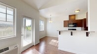This thoughtfully designed one-bedroom, one-bathroom residence offers a functional and modern single-floor layout. The galley-style kitchen is a standout feature, outfitted with rich dark wood cabinetry, granite countertops, black stainless steel appliances including a range, microwave, refrigerator, and dishwasher, all set atop warm hardwood-style flooring. Adjacent to the kitchen, a dedicated dining nook benefits from a large window and a pendant light fixture, creating a bright and inviting space for meals. The open living and dining area continues the hardwood-style flooring and is bathed in natural light from multiple windows and a glass door that offer views of surrounding greenery. A hallway connects the main living area to the private quarters. The bedroom is a comfortable retreat featuring plush carpeting, a generously sized closet, and a window with outdoor views. The bathroom is appointed with tile flooring, dark wood vanity cabinetry, and granite countertops, and connects to a walk-in closet with arched ceiling detail and a dedicated hanging rod. Clean white walls and neutral finishes throughout lend the space a cohesive, contemporary aesthetic that is both practical and welcoming.