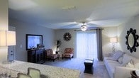 This single-story residence features a spacious main living area finished with wall-to-wall carpeting that adds warmth and comfort underfoot. The room is anchored by a ceiling fan with an integrated light fixture, providing both air circulation and ambient lighting. A large sliding glass door fitted with vertical blinds serves as the focal point of the exterior wall, allowing natural light to fill the space while offering access to the outdoors. Draping curtain panels frame the sliding door, adding a decorative touch. The living area offers generous room for seating arrangements, as evidenced by the accent chairs and additional furnishings placed throughout. A decorative sunburst wall accent adds visual interest to the neutral-toned walls. The layout flows naturally into a dining area, where a granite-style surface table is visible in the foreground, suggesting an open-concept design between living and dining spaces. A media console with a large mirror adds depth and dimension along one wall. The overall ambiance is bright, casual, and functional, with textured ceilings and neutral wall tones contributing to a clean, versatile interior aesthetic.
