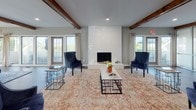 Located at 300 Hickory Hollow Pl in Nashville, TN, this single-floor community clubhouse offers a thoughtfully designed interior that blends modern and transitional styles. Rich hardwood-style flooring runs throughout the space, complemented by warm gray walls and an inviting, open layout.

The main lounge area is anchored by a striking white subway-tiled fireplace flanked by navy blue accent chairs and low-profile coffee tables. Exposed dark wood ceiling beams add architectural character and warmth overhead, while large windows and glass-paneled doors flood the room with natural light and provide views of the outdoor pool area.

An adjacent dining area features a glass-top table with upholstered burnt-orange chairs and a decorative area rug, creating an intimate yet functional gathering space. A bar-height counter with stylish stools serves as a transition zone, ideal for casual seating or socializing. Frosted glass sliding partitions offer the flexibility to divide areas as needed, adding a sleek, contemporary touch.

Decorative consoles, curated artwork, and layered area rugs contribute to the refined ambiance throughout. Multiple exterior access points connect seamlessly to the surrounding outdoor amenities, making this clubhouse a welcoming and well-appointed hub for community life.