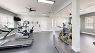 Located at Walden Creek Way in Greenville, South Carolina, this community fitness center offers a thoughtfully designed workout environment within a generously sized, single-floor space. The gym features a clean, open layout with durable rubber flooring suited for a wide range of exercise activities.

The facility is well-lit by a combination of recessed fluorescent lighting and an abundance of natural light streaming through multiple large, white-trimmed windows that line the sage-green accent walls. Ceiling fans are installed throughout to maintain comfortable airflow during workouts.

The cardio section is equipped with treadmills and a stair climber, complemented by wall-mounted televisions for entertainment during exercise. The strength training area includes a multi-station cable machine, spin bikes, a recumbent bike, a full dumbbell rack with a flat bench, and a SportsArt step platform. A large wall mirror enhances the sense of space and allows users to monitor their form. Stability and medicine balls are also available for functional training.

Structural columns punctuate the open floor plan while maintaining an airy, uncluttered feel. The light gray and sage-green color palette, white trim work, and bright natural light create a clean, welcoming atmosphere that encourages an active lifestyle.