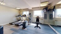 Located at 1530 Beacon St in Brookline, Massachusetts, this fitness center offers a well-equipped exercise facility designed to support a comprehensive workout experience. The space is organized across a single floor and is accessible via a carpeted corridor that connects to the main exercise area.

The fitness floor features durable, light-toned vinyl plank flooring and is divided into distinct functional zones. The cardio area is equipped with multiple treadmills, an elliptical trainer, and a stationary bike, all positioned near windows that allow natural light and views of the surrounding Brookline skyline. A wall-mounted flat-screen television provides entertainment during workouts.

The strength training area is generously outfitted with a multi-function cable and weight machine, adjustable benches, a dumbbell rack with a full range of free weights, and a stability ball, catering to a variety of exercise routines. Full-length mirrors line the walls, visually expanding the space and assisting with form and technique.

Practical amenities include a hand sanitizer dispenser, posted workout guides, and a rubber exercise mat. The corridor leading to the fitness center features patterned carpeting, crown molding, and overhead lighting, providing a clean and organized transition into the facility. Overall, the space is functional, well-maintained, and thoughtfully arranged for residents seeking convenient fitness access.