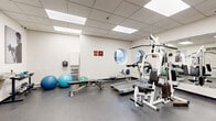 Located at 1241 Kilborn Place in Ottawa, Ontario, this fitness facility offers a comprehensive workout environment within a single, efficiently organized floor. The space features a drop-panel ceiling with recessed fluorescent lighting that ensures bright, even illumination throughout. A distinctive circular porthole window on one wall admits natural light and provides a view of the surrounding neighbourhood, while a large wall-mounted mirror visually expands the room and supports proper form during exercise.

The gym is well-equipped for a full-body workout. Cardio options include a treadmill, elliptical trainer, rowing machine, and two stationary bikes — one upright and one recumbent. Strength training is supported by a multi-function cable and weight machine, an adjustable Hoist workout bench, and a rack of dumbbells in varying sizes. Exercise balls in multiple sizes round out the functional fitness accessories.

The durable, speckled rubber flooring provides both safety and comfort underfoot, and the neutral white walls are accented by motivational black-and-white athletic photography. A wall-mounted water fountain with a bottle-filling station ensures convenient hydration. The space also includes a storage cabinet and wall-mounted equipment panels. The layout is thoughtfully arranged to allow freedom of movement across all exercise zones.