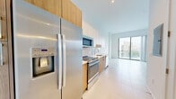 Located at 999 NW Seventh St in Miami, FL, this penthouse unit offers a thoughtfully designed layout with a clean, contemporary aesthetic. The open-plan kitchen is a standout feature, outfitted with stainless steel appliances including a side-by-side refrigerator with ice and water dispenser, a built-in microwave, an electric range, and a dishwasher. Warm wood-toned lower cabinetry is paired with crisp white upper cabinets, complemented by a light subway tile backsplash and a white quartz countertop with an undermount sink. Large-format ceramic tiles flow seamlessly throughout the space, enhancing the bright, airy feel. Floor-to-ceiling sliding glass doors flood the living area with natural light and provide direct access to a shared outdoor balcony, which features a distinctive perforated metal railing and open views of the surrounding Miami skyline. The bathroom is finished with matching tile flooring, a wood-cabinet vanity topped with a white countertop and framed mirror, and a glass-enclosed shower. A hallway with an electrical panel connects the main areas efficiently. The overall ambiance is modern, minimal, and light-filled, reflecting a distinctly urban Miami style.