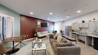 Located at 2274 5th Ave in Marion, Iowa, this single-floor combined kitchen and living room offers a thoughtfully designed and functional layout. The living area is anchored by a bold deep-red accent wall, against which a wall-mounted flat-screen television is flanked by contemporary artwork. A generously sized cream sofa with accent pillows faces the feature wall, complemented by paired round ottomans and a striped area rug that defines the seating zone. A second seating nook features a small round bistro table with modern chairs set against a muted sage-green accent wall, adding visual variety to the space. Double doors with frosted glass panels provide entry, maintaining an open and airy feel, while light wood-look flooring runs seamlessly throughout. The kitchen occupies a dedicated wall lined with grey shaker-style cabinetry, granite-look countertops, and a classic subway tile backsplash. Stainless steel appliances include a full-size refrigerator, dishwasher, and built-in microwave, all arranged along a practical galley-style run with an integrated sink. Recessed lighting throughout ensures bright, even illumination across the entire space, and a window allows natural light to filter into the dining and kitchen areas, enhancing the overall welcoming ambiance.