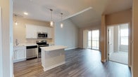 This single-floor residence offers a well-designed open-concept layout that seamlessly connects the kitchen, dining, and living areas. The kitchen is outfitted with white shaker-style cabinetry, light countertops, a full suite of stainless steel appliances—including a refrigerator, range, microwave, and dishwasher—and a central island that provides additional prep and seating space. Pendant lighting above the island adds a contemporary touch. The living area benefits from vaulted ceilings that enhance the sense of openness, and sliding glass doors lead directly to a private balcony with outdoor views, bringing in abundant natural light. Rich-toned hardwood-style flooring runs throughout the main living spaces, creating a cohesive and polished aesthetic. The bedroom is carpeted for comfort and features a ceiling fan, a large window with outdoor views, and generous closet access. The bathroom is finished with white cabinetry, a single vanity with a light countertop, a mirror, and hardwood-style flooring that continues the design language of the unit. Overall, the space presents a clean, modern ambiance with neutral tones and thoughtful finishes.
