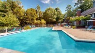 Located at 3550 Pleasant Hill Rd in Duluth, Georgia, this outdoor pool complex offers a well-appointed aquatic retreat set against a backdrop of mature, towering trees. The facility features multiple distinct pool areas, including a large, expansive swimming pool with crystal-clear blue water ideal for recreation and lap swimming, as well as a separate, smaller pool accented with striking cobalt blue tile detailing along its raised wall — a notable architectural feature that adds character to the space.

The pool decks are finished with a combination of paved brick and concrete surfaces, providing ample room for lounging and socializing. Teal-colored lounge chairs and sun umbrellas are thoughtfully arranged throughout the deck areas, offering comfort and shade for guests. A fenced perimeter ensures safety and privacy throughout the complex.

A pool house structure, clad in brick with a distinct pitched roofline, is situated adjacent to the pool area and serves as a functional support facility. The surrounding landscape is lush and green, with tall pines and deciduous trees framing the space and creating a natural, serene atmosphere. The overall ambiance is that of a well-maintained, resort-style outdoor amenity designed for relaxation and enjoyment in a community setting.
