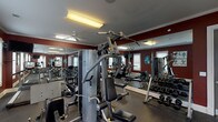 This fitness center is designed to support a comprehensive workout experience within a single, efficiently arranged space. The room features bold deep-red accent walls trimmed with white crown molding, rubber flooring for safety and durability, and large mirrored walls that visually expand the space while allowing users to monitor their form. Ceiling fans and fluorescent overhead lighting ensure the environment remains comfortable and well-lit throughout. Natural light enters through multiple white-trimmed windows fitted with blinds, adding brightness to the interior.

The cardio section includes treadmills, elliptical machines, and a stationary bike, several equipped with digital displays. A mounted flat-screen television provides entertainment during workouts. For strength training, the facility offers a multi-station cable machine, a dedicated dumbbell rack with a full range of free weights, a weight bench, and additional resistance equipment. Fitness reference charts are displayed on the walls to assist users with exercise techniques.

Entry is provided through a glass-paned door, and a fire extinguisher is conveniently mounted near the entrance. The overall layout is organized to allow users to move freely between equipment zones, making the fitness center a functional and practical amenity.