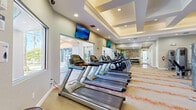 Located at 2450 Lake Debra Dr in Orlando, FL, this fitness center offers a comprehensive workout facility across two exercise rooms. The main exercise room is a generously proportioned space distinguished by elegant coffered and tray ceilings with recessed lighting that create a bright, polished atmosphere. Patterned carpet with bold orange accents adds a dynamic energy to the floor throughout. This primary area is lined with a full row of Matrix treadmills and elliptical trainers along one side, complemented by multiple cable and strength training machines, a functional trainer, a power rack with a punching bag, and a dumbbell rack. Wall-mounted flat-screen TVs provide entertainment throughout, and large windows and glass doors allow natural light to flow in while offering views of the outdoor surroundings. A motivational fitness-themed word art mural serves as a focal point on one wall. The secondary exercise room is a dedicated cycling studio featuring rows of spin bikes, a wall-mounted TV, and a vaulted ceiling with a large arched window that fills the room with natural light. Together, both rooms provide a well-rounded and inviting environment suited for a wide range of fitness activities.