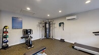 This fitness center at 13330 Blanco Rd in San Antonio, Texas, is a fully outfitted, single-floor exercise facility designed to support a wide range of workout routines. The space features carpet flooring throughout and is illuminated by recessed ceiling lighting, creating a bright and functional environment. Climate comfort is ensured by a wall-mounted mini-split air conditioning unit.

The cardio section is equipped with treadmills, an elliptical trainer, and a stationary recumbent bike, positioned near large windows that allow natural light to filter in and provide views of the surrounding greenery. A flat workout bench and step platforms are available for strength and conditioning exercises.

A centerpiece multi-functional cable machine offers versatile resistance training options, complemented by resistance bands and suspension straps mounted on the wall. A pull-up and cable station further expands the strength training capabilities of the space. Full-length wall mirrors enhance the sense of space and allow users to monitor their form during workouts.

The facility also includes a small amenity counter, a waste bin, and posted fitness center rules and guidelines, reflecting a community-oriented, well-maintained environment. A glass-paned exterior door provides convenient access and connects the space to the outdoors.