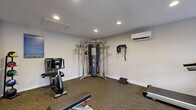This fitness center at 13330 Blanco Rd in San Antonio, Texas, is a fully outfitted, single-floor exercise facility designed to support a wide range of workout routines. The space features carpet flooring throughout and is illuminated by recessed ceiling lighting, creating a bright and functional environment. Climate comfort is ensured by a wall-mounted mini-split air conditioning unit.

The cardio section is equipped with treadmills, an elliptical trainer, and a stationary recumbent bike, positioned near large windows that allow natural light to filter in and provide views of the surrounding greenery. A flat workout bench and step platforms are available for strength and conditioning exercises.

A centerpiece multi-functional cable machine offers versatile resistance training options, complemented by resistance bands and suspension straps mounted on the wall. A pull-up and cable station further expands the strength training capabilities of the space. Full-length wall mirrors enhance the sense of space and allow users to monitor their form during workouts.

The facility also includes a small amenity counter, a waste bin, and posted fitness center rules and guidelines, reflecting a community-oriented, well-maintained environment. A glass-paned exterior door provides convenient access and connects the space to the outdoors.