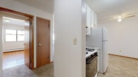 Located at 3612 Lynn Ave in St. Louis Park, MN, this two-bedroom, one-bathroom unit offers a practical and well-organized single-floor layout. The galley-style kitchen is thoughtfully appointed with white shaker cabinetry, marble-look countertops, a mosaic tile backsplash, tile flooring, and white appliances including a gas range and full-size refrigerator — providing efficient use of space for everyday cooking. Adjacent to the kitchen, a carpeted hallway connects the main living areas, with warm wood-toned doors adding a classic touch throughout. The living area features neutral carpet, white walls, and a window with a baseboard heating unit, keeping the space bright and functional. Both bedrooms are highlighted by rich hardwood flooring that adds warmth and character. The primary bedroom includes louvered bi-fold closet doors and a window offering natural light, while a baseboard heater ensures year-round comfort. The full bathroom is clean and updated, featuring a white vanity with ample drawer storage, solid-surface countertop, brushed nickel fixtures, and a tiled shower visible in the mirror. The unit's straightforward layout and mix of hardwood and carpet flooring make it a practical and comfortable home.