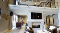 This two-story space combines contemporary design with functional elegance across two well-appointed levels. The main floor centers around a dramatic living area anchored by a floor-to-ceiling textured white fireplace surround, complete with a linear hearth and a large wall-mounted television. Double-height ceilings create an airy, open atmosphere, while large floor-to-ceiling windows invite abundant natural light and views of the surrounding exterior. Soft, neutral flooring and a curated mix of leather seating and layered area rugs contribute to a refined yet welcoming ambiance.

The upper level features a mezzanine overlooking the main floor, framed by sleek glass-panel railings with cable accents. This elevated space hosts a versatile area with a dark wood conference-style dining table paired with leather and gold-legged chairs, illuminated by geometric brass pendant lights suspended from a coffered ceiling with blue-grey insets. An adjacent built-in workstation with dark granite surfaces and task lighting adds a functional dimension. The overall design language throughout is modern and polished, blending bold architectural details with warm, sophisticated finishes.