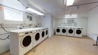 This communal laundry facility is designed for efficiency and convenience, offering a well-organized space equipped with a substantial number of front-loading washing machines and dryers. The machines are arranged along two walls, maximizing the use of the available floor space and allowing multiple users to do laundry simultaneously. The room features durable, light-toned tile flooring that is easy to maintain and clean. Overhead fluorescent lighting runs the length of the ceiling, ensuring the space is brightly and evenly illuminated throughout. Exposed utility pipes and conduits are routed neatly along the white ceiling and walls, reflecting a practical, no-frills infrastructure. A utility sink and additional plumbing fixtures are visible along one wall, adding to the room's functionality. The white walls and ceiling contribute to a clean, bright atmosphere. The layout is straightforward and accessible, providing ample room to move between machines. Overall, this laundry facility prioritizes practicality, cleanliness, and capacity, making it a highly functional shared amenity.