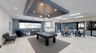 This contemporary amenity space is thoughtfully designed to offer residents or guests a versatile environment for recreation, relaxation, and socializing. The open-plan layout centers around a full-size billiards table, anchored by a neutral area rug and illuminated by a striking geometric pendant light fixture suspended from a bold, recessed tray ceiling finished in deep slate gray — a standout architectural detail that adds visual depth to the space.

To one side, a comfortable lounge area features plush seating arranged around a large-format projection screen, ideal for movie nights or group gatherings. Large windows along the lounge wall allow natural light to fill the space while offering views of the surrounding outdoors. On the opposite side, a dining area with wooden tables and upholstered blue chairs provides a casual setting for meals or casual meetings.

Light wood-tone flooring runs throughout, complementing the neutral and cool-toned color palette of grays, blues, and whites. Glass-paneled walls provide a seamless visual connection to an adjacent fitness center, where exercise equipment is visible, reinforcing the space's focus on active living. Recessed lighting and modern finishes throughout contribute to a clean, polished ambiance that balances functionality with contemporary style.