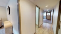 This single-floor space is defined by a well-proportioned hallway that serves as the central artery connecting its various rooms. The corridor is finished with light-toned hardwood-style flooring that runs continuously throughout, creating a cohesive and easy-to-maintain surface underfoot. The walls are painted in a soft, neutral cream tone, contributing to a clean and understated aesthetic that keeps the space feeling bright and open.

A notable design detail is the use of gold-toned door and wall trims, which add a subtle warmth and visual interest against the pale walls. Doorframes along the hallway are fitted with this consistent gold trim, lending a degree of polish to the overall interior. Recessed ceiling lighting provides even illumination throughout the corridor.

At the far end of the hallway, a window allows natural light to filter in, offering a glimpse of an exterior balcony or entryway beyond. This endpoint opens into a carpeted area, suggesting a transition into a living or lounge space. The overall ambiance of the space is practical and tidy, with a neutral palette and consistent finishes that create a calm, functional environment suitable for a variety of uses.
