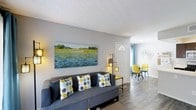 This single-floor space offers a welcoming and well-appointed interior designed with a modern, contemporary aesthetic. The living area serves as the heart of the home, centered around a plush gray sofa accented with bold yellow and blue polka-dot throw pillows. A large landscape painting adorns the light gray wall, adding a splash of color and artistic character to the room. Warm ambient lighting from a stylish floor lamp complements the natural light filtering through the front entryway, which features a classic white door with a distinctive sunburst transom window above. The flooring throughout is a clean, wood-look plank finish that adds warmth and continuity across the open layout. Transitioning from the living area, a casual dining nook is visible toward the rear of the space, furnished with vibrant yellow chairs that echo the home's cheerful color story. Decorative wall art in a honeycomb motif adds a modern touch to the dining area. Blue curtains frame the windows, tying together the cohesive color scheme. The textured ceiling and neutral wall tones provide a versatile backdrop, making the space feel both lively and effortlessly put-together throughout.