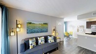 This single-floor space offers a welcoming and well-appointed interior designed with a modern, contemporary aesthetic. The living area serves as the heart of the home, centered around a plush gray sofa accented with bold yellow and blue polka-dot throw pillows. A large landscape painting adorns the light gray wall, adding a splash of color and artistic character to the room. Warm ambient lighting from a stylish floor lamp complements the natural light filtering through the front entryway, which features a classic white door with a distinctive sunburst transom window above. The flooring throughout is a clean, wood-look plank finish that adds warmth and continuity across the open layout. Transitioning from the living area, a casual dining nook is visible toward the rear of the space, furnished with vibrant yellow chairs that echo the home's cheerful color story. Decorative wall art in a honeycomb motif adds a modern touch to the dining area. Blue curtains frame the windows, tying together the cohesive color scheme. The textured ceiling and neutral wall tones provide a versatile backdrop, making the space feel both lively and effortlessly put-together throughout.