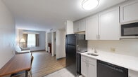 Located at 835 Upper Union St in Franklin, MA, this single-floor suite offers a thoughtfully designed layout accommodating three bedrooms and three full bathrooms. The open entryway leads into a central corridor connecting all areas of the suite, with light hardwood-style flooring running throughout and a clean, neutral color palette that creates a bright, cohesive atmosphere.

The living area is simply furnished with a comfortable sofa and features a window dressed with layered curtains that allow natural light to filter in. Adjacent to the living space, a compact dining table with chairs provides a convenient area for meals. A refrigerator and additional storage are accessible nearby, adding practical functionality.

Each of the three bedrooms follows a consistent, modern design, featuring dual beds dressed in crisp white linens with navy blue accents, set against a bold warm-toned wood headboard panel that serves as a striking focal point. Light wood flooring completes the clean, welcoming aesthetic. Windows with draping curtains allow for natural light and privacy.

The three bathrooms are finished with large-format light tile flooring and include dark-toned vanities topped with white countertops and backlit mirrors. Each bathroom features a glass-enclosed shower stall, offering a sleek, contemporary look. One bathroom is notably spacious and ADA-accessible, with grab bars and an open floor plan for ease of movement throughout.