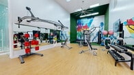 Located at 100 Avalon Gardens Dr in Nanuet, NY, this comprehensive fitness and recreation facility spans a generous single floor, offering residents a wide range of athletic amenities under one roof. The main fitness area is a standout space, boasting soaring double-height ceilings, large arched windows that flood the room with natural light, gleaming hardwood-style flooring, and full-wall mirrors that amplify the sense of scale. It is well-equipped with cardio machines including treadmills and stationary bikes, a full dumbbell rack, and ample open floor space for stretching and functional training. A dedicated fitness studio features vaulted ceilings, a vibrant geometric accent wall, wall-to-wall mirrors, a ceiling-mounted large industrial fan, and a variety of strength training equipment including a multi-station cable machine, boxing bags, and a wall-mounted rack system. The indoor basketball court is a spacious, bright area with a teal and blue sport-surface floor, regulation markings, a wall-mounted adjustable backboard, and natural light from surrounding windows. The racquetball court is fully enclosed with pristine white walls, hardwood flooring with red court markings, and overhead skylight-style lighting. Glass partition walls connect several zones, creating visual openness throughout the facility.