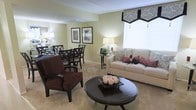 This two-floor residence offers a thoughtfully designed interior centered around an open-concept living and dining area. The living space is anchored by a cream upholstered sofa accented with floral and pink throw pillows, complemented by a rich dark-wood round coffee table and a deep burgundy armchair, creating a warm and inviting seating arrangement. Soft, neutral carpeting runs throughout, adding to the overall comfort of the space. A large window dressed with a decorative black-and-white valance allows generous natural light to fill the room. Adjacent to the living area, the dining space accommodates a dark wood table and matching chairs, styled beneath framed botanical wall art that adds a touch of color to the light green walls. Warm ambient lighting from table lamps enhances the cozy atmosphere throughout. The cohesive color palette of soft greens, creams, and dark wood tones ties both areas together seamlessly, resulting in a space that feels both elegant and livable across its two floors.
