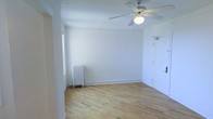 This single-floor space presents a clean, minimalist aesthetic defined by its bright white walls and warm-toned hardwood floors that run throughout. The main living area is well-proportioned, offering an open and airy feel that is enhanced by natural light filtering in through a side window. A classic ceiling fan with an integrated light fixture is centrally mounted, providing both ventilation and ambient lighting to the room. A traditional cast iron radiator sits along one wall, serving as both a functional heating element and a charming architectural detail that hints at the building's character. Crown molding lines the upper perimeter of the room, adding a subtle decorative finish to the otherwise understated interior. The entry door is painted white and features standard hardware, contributing to the cohesive, neutral palette found throughout the space. The overall ambiance is light, clean, and versatile, making it well-suited for a variety of uses. The uncluttered design and hardwood flooring provide a solid foundation for customization, while the classic architectural details lend the space a timeless quality.