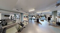Located at 35 Folly Road Blvd in Charleston, South Carolina, this single-floor fitness center offers a well-appointed and comprehensive workout facility. The main gym area is generously sized, featuring a wide selection of cardio equipment including treadmills, elliptical trainers, and stair steppers, alongside a multi-station strength training machine and a dedicated free weights section complete with a full dumbbell rack and adjustable benches. Large windows line the perimeter, inviting abundant natural light and offering views of the surrounding greenery, while ceiling fans and overhead fluorescent lighting ensure a comfortable environment. Full-length mirrors along the walls enhance the sense of space and allow exercisers to monitor their form. The flooring transitions thoughtfully between durable, easy-clean surfaces in the main gym area and light hardwood in the studio. A separate studio room features hardwood flooring, floor-to-ceiling mirrors, a ballet barre, stability balls, kettlebells, and stationary bikes — ideal for group classes or individual training sessions. A sliding barn door connects the two areas, and glass-paneled entry doors provide easy access. The space is clean, bright, and thoughtfully organized, making it a functional and inviting facility for a wide range of fitness activities.