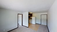 This single-floor space offers a straightforward, functional layout that connects the living area directly to the kitchen. The living area is finished with neutral wall-to-wall carpet and features dark brown door trim that provides contrast against the light-colored walls. A textured popcorn ceiling runs throughout, adding a classic residential feel. Two interior doors are visible from the main living space, offering access to additional rooms within the unit.

The kitchen area is defined by tile flooring, transitioning clearly from the carpeted living space. It is equipped with essential appliances including a white electric range and a full-size white refrigerator. Dark-stained cabinetry lines the upper wall, providing ample storage. An overhead light fixture illuminates the kitchen area. The layout allows for easy movement between the living and kitchen zones, making the space practical for everyday use. An exterior door situated near the kitchen provides a secondary point of entry. Overall, the space presents a utilitarian design with straightforward finishes suited for everyday living.