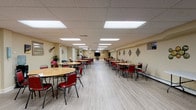 This single-floor venue is designed to accommodate group gatherings and events with a well-organized, functional layout. The centerpiece of the space is a generously sized main hall featuring light wood-tone luxury vinyl flooring, a drop tile ceiling with recessed panel lighting, and warm beige walls. Multiple round tables paired with red upholstered chairs are arranged throughout, offering comfortable seating for a sizable number of guests. Decorative wall accents, including diamond-shaped mirrors and guitar wall art, add character to the hall. A flat-screen television mounted at the far end enhances the space's versatility for presentations or entertainment.

Adjacent to the main hall is a fully equipped kitchen featuring light wood cabinetry, dark granite-style countertops, tile flooring, and black appliances including a gas range, microwave, and refrigerator — well-suited for catering or food preparation during events.

A separate lounge or breakout room offers a quieter retreat, finished with the same light vinyl flooring and warm tan walls. It includes a large flat-screen television, a leather armchair, and decorative wall pieces, creating a relaxed, comfortable atmosphere. Overall, the space is practical, versatile, and well-maintained.