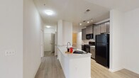 This single-floor residence is thoughtfully designed with a functional, open layout that connects the main living spaces seamlessly. The entryway leads down a well-lit hallway with wood-look flooring that flows throughout the common areas, lending a clean and cohesive aesthetic.

The kitchen serves as a central hub, featuring a peninsula with a light stone-patterned countertop and an undermount sink — ideal for casual dining or food preparation. Gray shaker-style cabinetry lines the walls, complemented by black appliances including a gas range, built-in microwave, and a full-size refrigerator. Track lighting above provides focused illumination throughout the cooking area.

The open living area is generously proportioned, with sliding glass doors fitted with vertical blinds that allow abundant natural light and provide access to an outdoor area. The space flows directly from the kitchen, creating an inviting, connected environment.

The bedroom offers plush carpeting, a large window with outdoor views, and access to a spacious walk-in closet with multi-level shelving. The full bathroom includes a soaking tub with shower, a wall-mounted sink, a lighted vanity mirror, and accessibility grab bars. A dedicated in-unit laundry area with side-by-side washer and dryer completes the space.