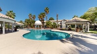 This single-level outdoor amenity space showcases a resort-inspired design centered around a generously sized freeform swimming pool with a smooth concrete surround ideal for lounging. The pool's curving shape and clear blue water create a relaxing focal point for the community. Adjacent to the main pool is a raised spa feature, elegantly bordered by natural stone rockwork and desert landscaping, adding a layered visual interest to the space.

A distinctive dome-topped gazebo with white columns stands poolside, serving as a decorative and functional landmark within the complex. Complementing it is a separate covered pavilion featuring a terracotta tile roof, open-air seating with bar-height tables and chairs, ceiling fans, and columned supports — offering a shaded retreat with direct views of the pool area.

The surrounding landscape is richly planted with mature palm trees, tropical shrubs, and manicured greenery, evoking a warm desert oasis atmosphere. The Mediterranean-style architecture of the adjacent buildings, characterized by stucco exteriors, tile rooflines, and arched windows, ties the entire space together cohesively. Under abundant natural sunlight and clear blue skies, this outdoor area provides an inviting and well-appointed setting for relaxation and recreation.