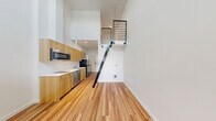 This 1-bedroom, 1-bathroom loft-style unit in Seattle, WA offers a thoughtfully designed layout that maximizes every inch of its compact footprint. The moment you enter, soaring double-height ceilings create a striking sense of verticality and openness that defies the unit's modest size. Natural light filters in through a clerestory window high on the wall and a standard window in the living area, keeping the space bright and airy throughout the day.

The open-plan kitchen and living area features warm-toned hardwood flooring that runs throughout the unit, grounding the crisp white walls and ceilings. The kitchen is efficiently arranged along one wall, equipped with light wood cabinetry, stainless steel appliances including a built-in microwave and refrigerator, and a clean countertop with an integrated sink.

A striking black metal staircase with horizontal rail detailing climbs to the lofted bedroom, which is tucked above and accessible via the upper landing. The bedroom is an intimate, cozy retreat with a window offering neighborhood views and a sliding closet.

The bathroom is clean and functional, featuring warm wood vanity cabinetry, a white countertop, a toilet, and a tub/shower combination with a frosted glass panel, all finished with consistent hardwood flooring. The overall aesthetic is modern, minimal, and efficiently designed.