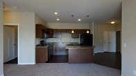 This single-floor space showcases an open-concept layout that connects the kitchen and living area in a cohesive, functional design. The kitchen is outfitted with rich, dark wood cabinetry that extends to the upper walls, providing ample storage alongside a full suite of black appliances, including a refrigerator, range, and built-in microwave. A double sink is positioned beneath recessed lighting, while two warm-toned pendant lights hang above the standout curved breakfast bar, which serves as both a functional workspace and a natural dividing element between the kitchen and the adjoining living space. The countertops feature a neutral stone-toned surface that complements the dark cabinetry throughout. The kitchen floor is finished in dark hardwood-style flooring, which transitions into plush, neutral-toned carpeting in the living area, creating a clear and inviting distinction between the two zones. Recessed ceiling lights illuminate the kitchen efficiently, contributing to a warm and practical ambiance. A doorway visible at the rear of the space suggests additional rooms beyond, hinting at a broader, well-connected floor plan throughout the home.