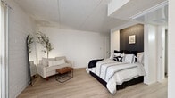 Located at 1030 S Dobson Rd in Mesa, Arizona, this efficiently designed one-bedroom, one-bathroom unit offers a thoughtfully utilized single-floor layout. The generously proportioned bedroom serves as the centerpiece of the space, featuring light wood laminate flooring, white painted brick-style accent walls, and a striking dark slatted wood feature wall behind the bed. A sitting area with a neutral-toned sofa, leather ottoman, and decorative greenery adds versatility to the room. Large vertical blinds over a sliding glass door panel allow for natural light control while maintaining privacy. The open-concept design includes a dedicated workspace nook, creating a functional and flexible living environment. The galley-style kitchen is lined with warm wood cabinetry on both sides, offering ample storage, and is equipped with black appliances including a refrigerator and electric range, complemented by light countertops and wood-look flooring that carries throughout. The bathroom features a wood-vanity with a single sink, a full tub-shower combination with tile surround, and clean white finishes. A closet provides additional storage. The unit's cohesive black, white, and natural wood color palette creates a modern yet inviting ambiance throughout.