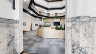 This space showcases a striking blend of historic architectural grandeur and modern interior design. The centerpiece is a spacious octagonal atrium rising dramatically to impressive heights, crowned by an encircling upper-level balcony with ornate black ironwork railings and deep black crown molding that contrasts sharply against crisp white walls. A skylight above the balcony level allows natural light to filter into the space.

The main floor features patterned grey carpet tile throughout, creating a neutral, professional foundation. A central reception-style station constructed of light wood panels is adorned with lush planters housing a variety of succulents and tall greenery, adding vibrant life to the interior.

Distinct functional zones are defined within the open floor plan, including a built-in nook with a fireplace, collaborative seating areas with colorful accent chairs, and casual work surfaces. Historic black-and-white photographs mounted along the walls reference the building's heritage.

The entryway retains original mosaic tile flooring and a formal black double-door vestibule, flanked by veined marble columns and a marble staircase. Additional private rooms with warm wood doors line the perimeter, accessible from the main atrium floor.