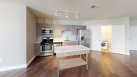 Located in the heart of downtown Rochester, NY, this single-floor residence offers a thoughtfully designed layout with modern finishes throughout. The open-concept living and kitchen area is anchored by rich hardwood flooring and track lighting, creating a bright, contemporary atmosphere. The kitchen is well-appointed with sleek wood-grain cabinetry, white quartz countertops, a white subway tile backsplash, and a full suite of stainless steel appliances including a gas range, built-in microwave, and French door refrigerator. A freestanding island provides additional prep and storage space, while an in-unit washer and dryer are conveniently tucked into a dedicated laundry closet nearby. The bedrooms feature plush wall-to-wall carpeting and generous windows offering striking views of the Rochester skyline, filling each room with natural light. The bathroom is finished with large-format tile flooring, a walnut-toned vanity with a white countertop, a framed mirror, and a glass-enclosed walk-in shower with full tile surround. Neutral tones and clean architectural lines carry throughout the space, lending a cohesive, move-in-ready aesthetic in a prime urban location.