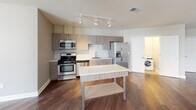 Located in the heart of downtown Rochester, NY, this single-floor residence offers a thoughtfully designed layout with modern finishes throughout. The open-concept living and kitchen area is anchored by rich hardwood flooring and track lighting, creating a bright, contemporary atmosphere. The kitchen is well-appointed with sleek wood-grain cabinetry, white quartz countertops, a white subway tile backsplash, and a full suite of stainless steel appliances including a gas range, built-in microwave, and French door refrigerator. A freestanding island provides additional prep and storage space, while an in-unit washer and dryer are conveniently tucked into a dedicated laundry closet nearby. The bedrooms feature plush wall-to-wall carpeting and generous windows offering striking views of the Rochester skyline, filling each room with natural light. The bathroom is finished with large-format tile flooring, a walnut-toned vanity with a white countertop, a framed mirror, and a glass-enclosed walk-in shower with full tile surround. Neutral tones and clean architectural lines carry throughout the space, lending a cohesive, move-in-ready aesthetic in a prime urban location.