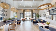 Located at 415 Curtis Pkwy SE in Calhoun, Georgia, this elegantly designed entrance and clubhouse space makes a striking first impression. The single-floor layout is unified by light-toned herringbone hardwood flooring that flows seamlessly throughout, complementing the bright, airy aesthetic of the interior.

Soaring coffered and beam-detailed ceilings lend architectural distinction to the space, while an array of woven rattan and gold-trimmed pendant lights provide warm, layered illumination. A bold geometric-patterned tile accent wall serves as a dramatic focal point, framing a built-in bar and counter area outfitted with high stools — ideal for casual collaboration or refreshments.

The lounge area features plush navy blue armchairs arranged around a low coffee table, anchored by a curated gallery wall of framed artwork displayed on shiplap paneling. Intimate bistro-style tables and leather chairs offer additional seating near large, floor-to-ceiling windows and glass doors that flood the space with natural light and provide direct views of an outdoor pool area.

The entryway welcomes visitors through black-framed glass double doors, accented by a decorative chandelier overhead and vibrant abstract artwork on the walls. Throughout, thoughtful décor including live plants, sculptural accessories, and colorful throw pillows adds warmth and personality to this refined, community-focused space.